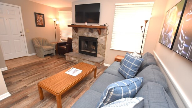 A living room with a stone fireplace, a flat-screen TV mounted above it, a grey sofa with blue and white patterned cushions, a wooden coffee table, and a striped armchair. - Condo at the University of Notre Dame D3 (South Bend)