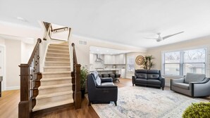 A spacious living area with a wooden staircase, a kitchen in the background, and a large window providing natural light. - Modern Home Just 10 Mins to Notre Dame 18384 (South Bend)