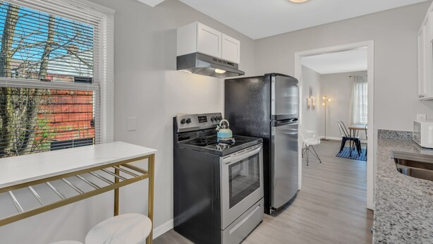 Une cuisine moderne dotée d’une cuisinière, d’un réfrigérateur et de meubles de rangement blancs, le tout en acier inoxydable. On y trouve une fenêtre avec des stores, un coin repas avec une table et des chaises, ainsi qu’un plan de travail avec un évier.