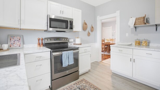A modern kitchen with white cabinets, a stainless steel oven, a microwave, and a coffee maker. There is a patterned rug on the floor and a wooden dining table in the background. - 3 Bedroom 2 Bath Home Near Notre Dame 123nd (South Bend)