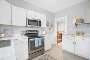 A modern kitchen with white cabinets, a stainless steel oven, a microwave, and a coffee maker. There is a patterned rug on the floor and a wooden dining table in the background. - 3 Bedroom 2 Bath Home Near Notre Dame 123nd (South Bend)