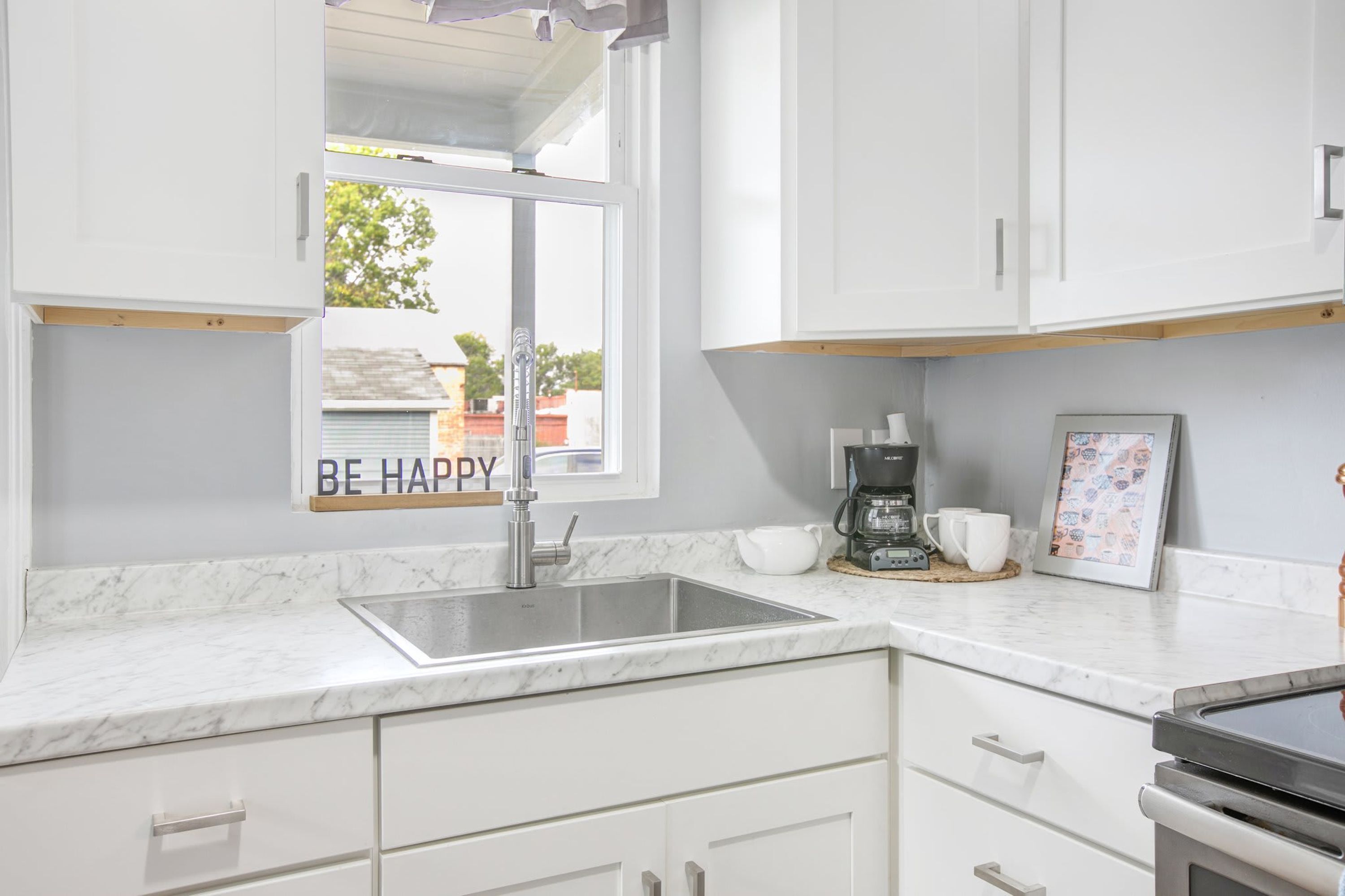 A modern kitchen with white cabinets, a marble countertop, a stainless steel sink, and a coffee maker.