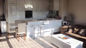 Una cocina moderna con armarios blancos y una isla central, un área de comedor con mesa y sillas blancas, y una sala de estar con sofá y mesa de centro.