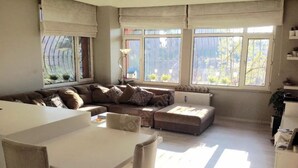 Una sala de estar con un sofá modular, una mesa de comedor con sillas y ventanas grandes con cortinas.