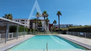 Una piscina rettangolare con un prendisole in legno di fronte, circondata da una recinzione metallica e palme.