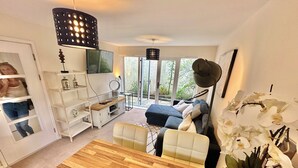 A modern living room with a wooden coffee table, a sofa, and a TV on a white stand. There is a large window with a view of greenery outside.