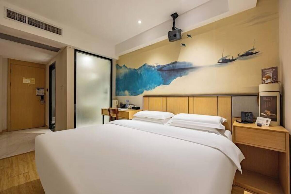 A hotel room with a bed, a desk, a chair, a nightstand, and a wall with a painting of ships.