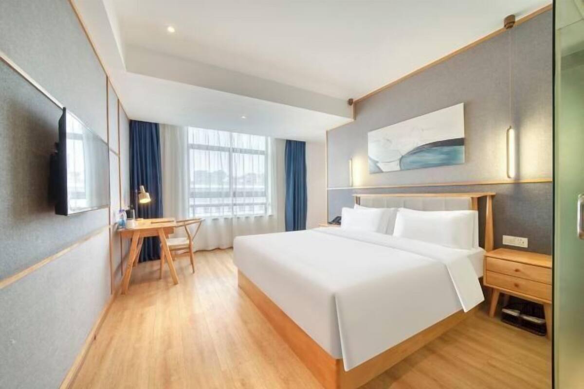 A hotel room with a bed, bedside table, desk, chair, and a large window with curtains.