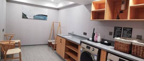 A compact laundry room with a washing machine, dryer, and a sink. There are wooden chairs and a small table, along with a shelf holding baskets and a bottle.