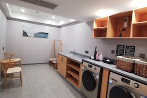 A compact laundry room with a washing machine, dryer, and a sink. There are wooden chairs and a small table, along with a shelf holding baskets and a bottle. - Greentree Eastern Hotel (Zhongshan West Road, Xiuzhou District, Jiaxing) (Jiaxing)