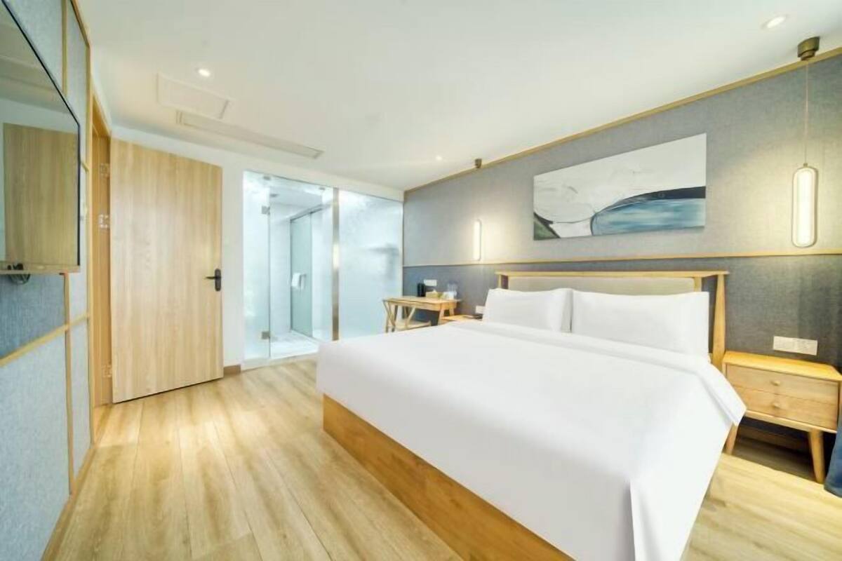 A modern hotel room with a large bed, wooden flooring, a glass door leading to a balcony, and a minimalist decor.