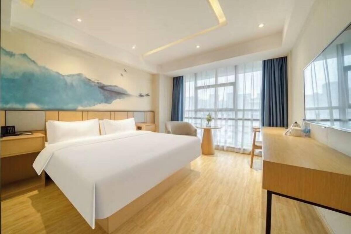A hotel room with a large bed, a desk, a chair, a nightstand, and a window with curtains.