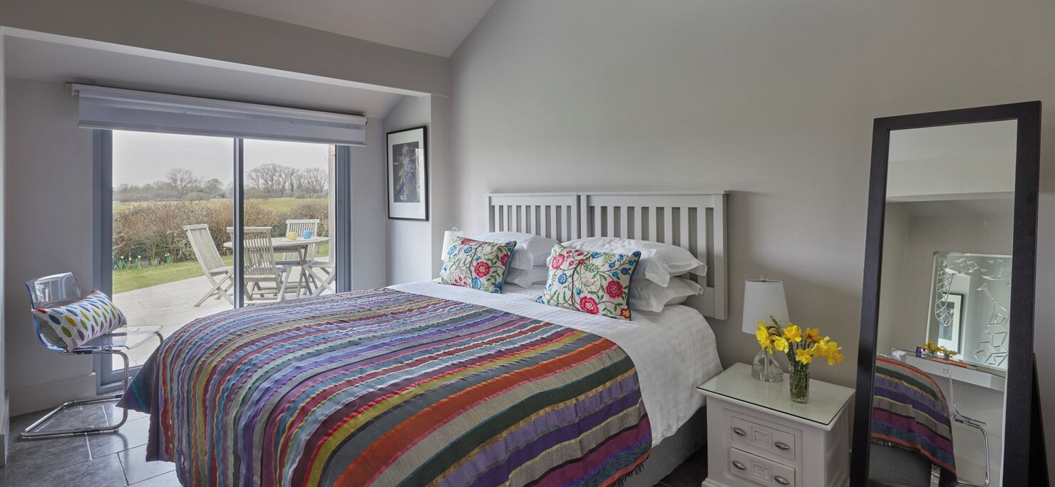 Une chambre dotée d’un grand lit, d’une table de chevet avec une lampe, d’une chaise et d’une porte-fenêtre coulissante donnant sur un espace extérieur.
