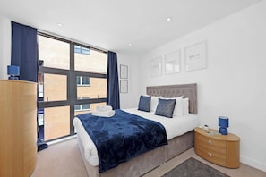Une chambre moderne avec une grande fenêtre, un lit recouvert d’une couverture bleu foncé, une commode en bois et une table de chevet avec une lampe.