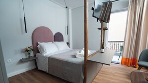 A modern bedroom with a bed, a TV mounted on a wooden frame, a small table, and a view of the balcony. - Modern & Sunny apartment with Garden View in the heart of the city center NYX 1 (Tripoli)