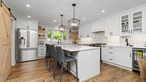 Une cuisine moderne avec des meubles blancs, un réfrigérateur en acier inoxydable, un îlot central doté de tabourets de bar, un éclairage suspendu et une vue sur la verdure à l’extérieur.