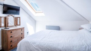 Quarto com teto inclinado, claraboia, cama com roupa de cama branca, cómoda de madeira com gavetas, televisão fixada na parede e espelho com moldura de madeira.