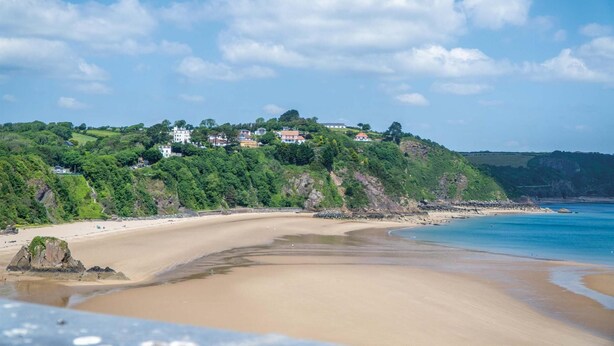 Paisagem costeira com praia de areia, afloramento rochoso e colina com edifícios.