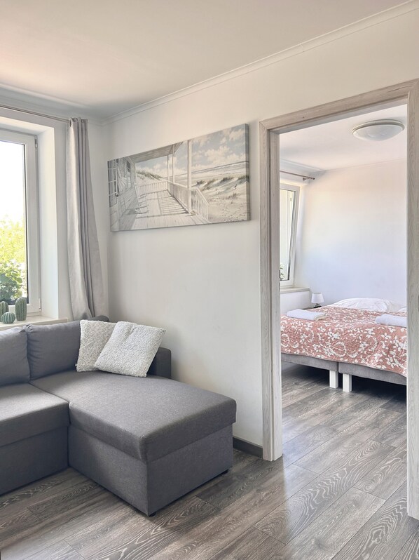 A modern bedroom with a grey sofa, a bed with a floral bedspread, and a painting of a beach scene. - Apartamenty Golf Dyndeby (Nexø)