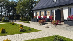 A modern building with a sloped roof, large windows, and a paved entrance area with potted plants and outdoor seating. - Apartamenty Golf Dyndeby (Nexø)