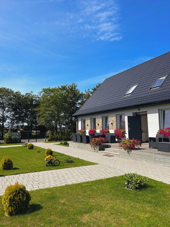 A modern building with a sloped roof, large windows, and a paved entrance area with potted plants and outdoor seating. - Apartamenty Golf Dyndeby (Nexø)