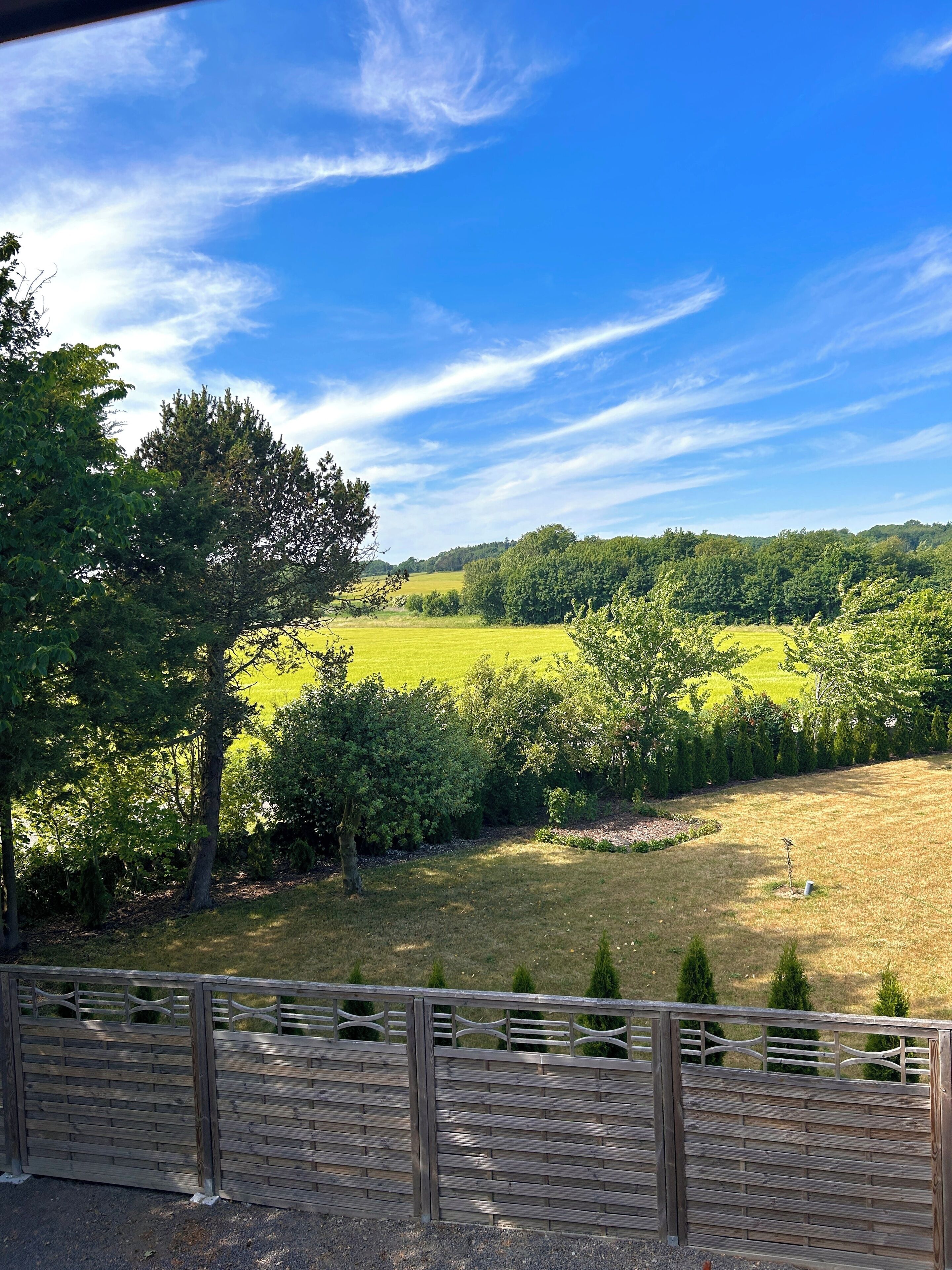 A view of a rural landscape with a wooden fence, trees, and a clear blue sky.