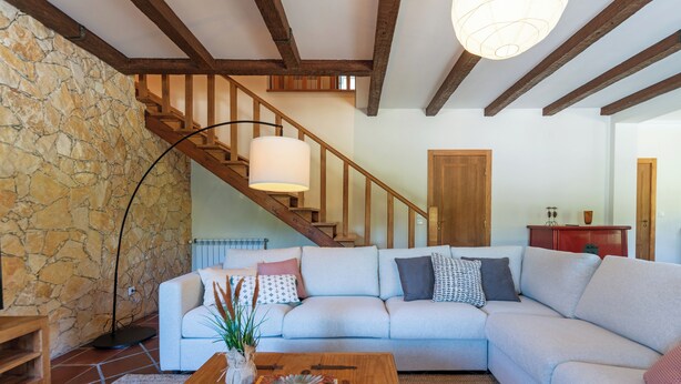 Un salon avec un mur en pierre, un escalier en bois, un canapé de couleur claire, une table basse en bois sur laquelle repose un livre, et un lampadaire.