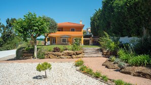 Une maison à deux étages, avec un toit recouvert de tuiles rouges, entourée d’un jardin aménagé où poussent différentes espèces de plantes, et un chemin de gravier menant à l’entrée.