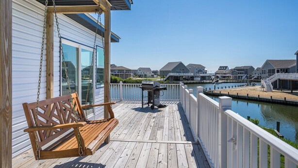 Une terrasse en bois avec une chaise suspendue et un barbecue, donnant sur un canal bordé de maisons.