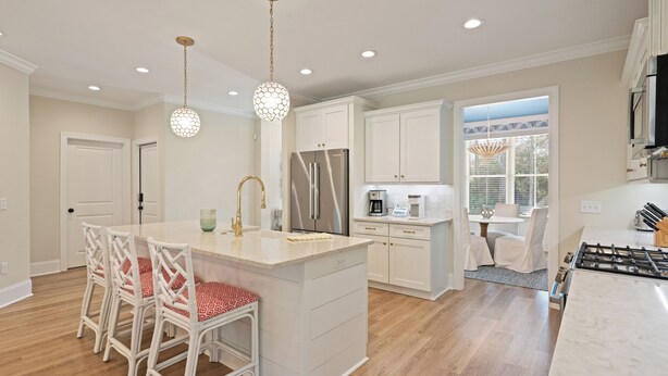 A modern kitchen with white cabinetry, a central island, and a dining area with wicker chairs. - Executive  style home with 4 bedrooms and 3.5 baths. (Saint Simons Island)
