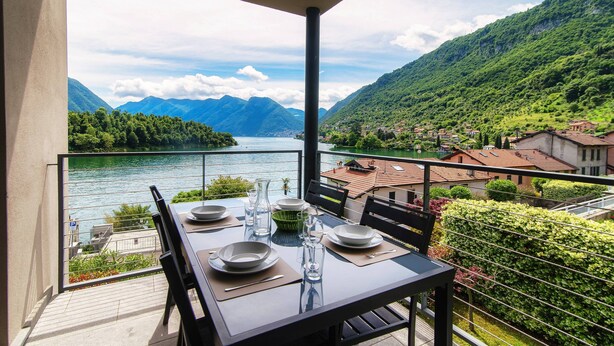 A balcony table set with plates, glasses, and cutlery, overlooking a lake and mountains. - Lake's Dream (Tremezzina)