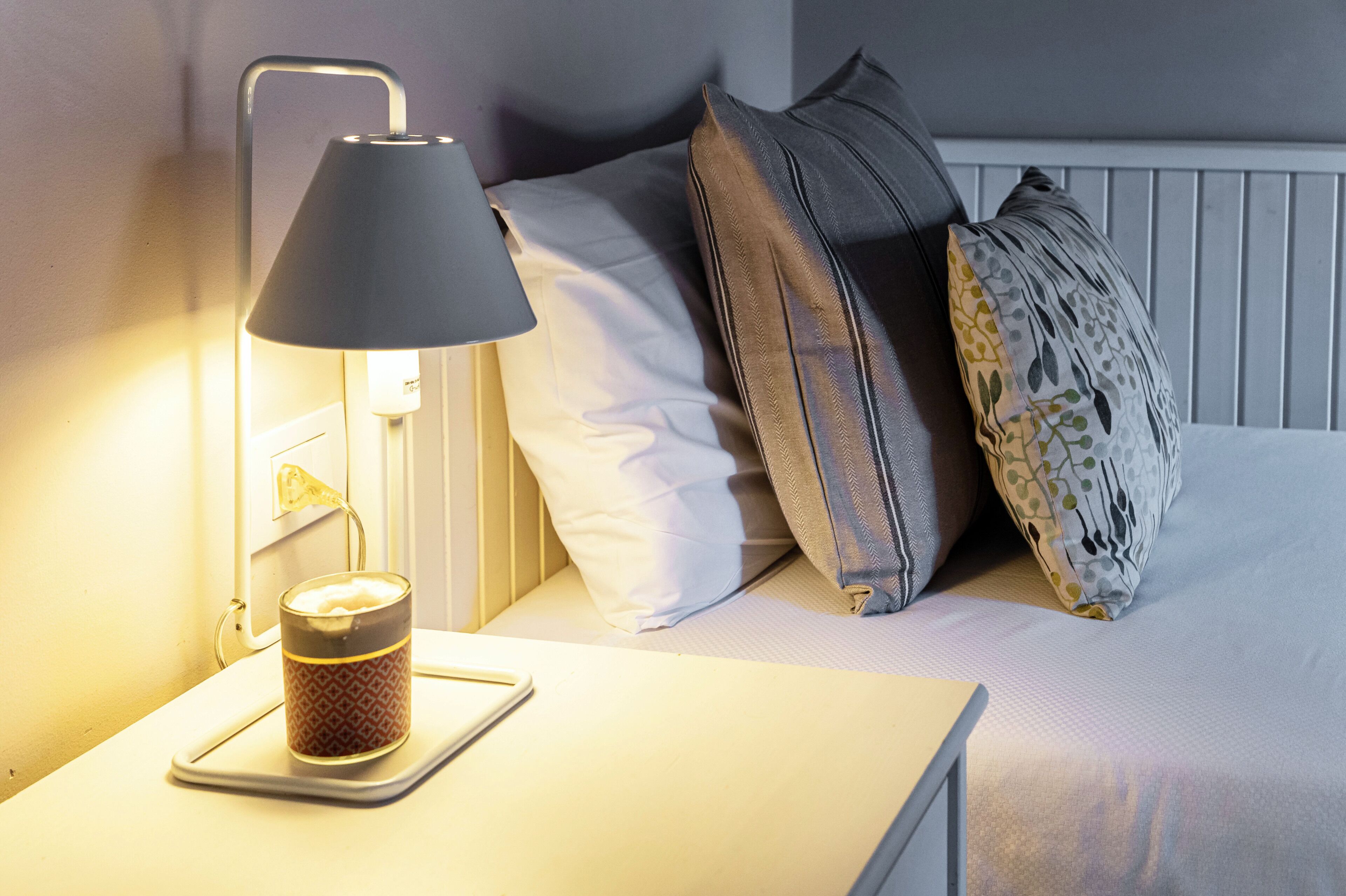 Une table de chevet avec une lampe et une bougie, un lit avec une literie blanche et plusieurs oreillers, et un luminaire fixé au mur.