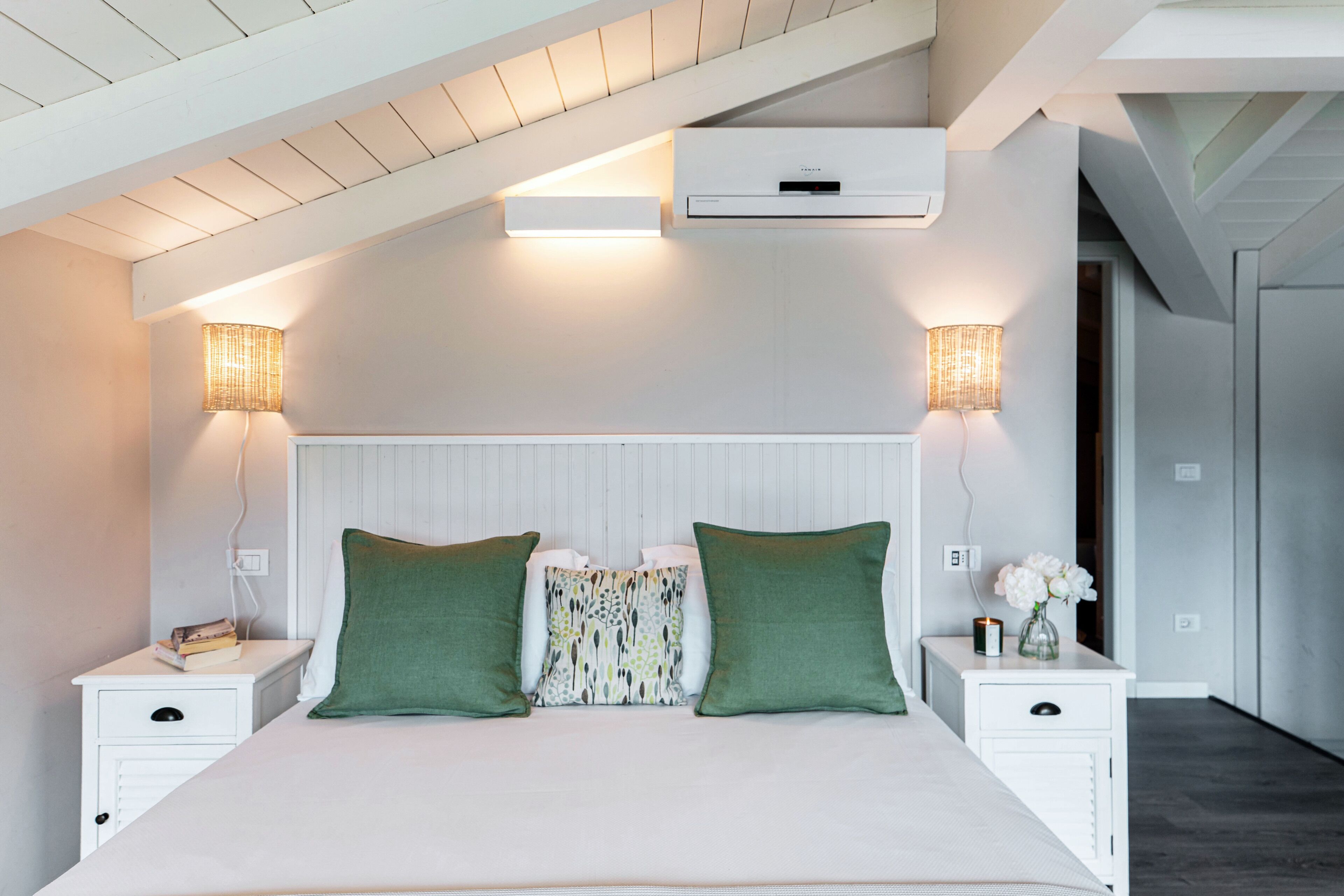A bedroom with a sloped ceiling, a bed with green pillows, two bedside tables with lamps, and a wall-mounted air conditioner.