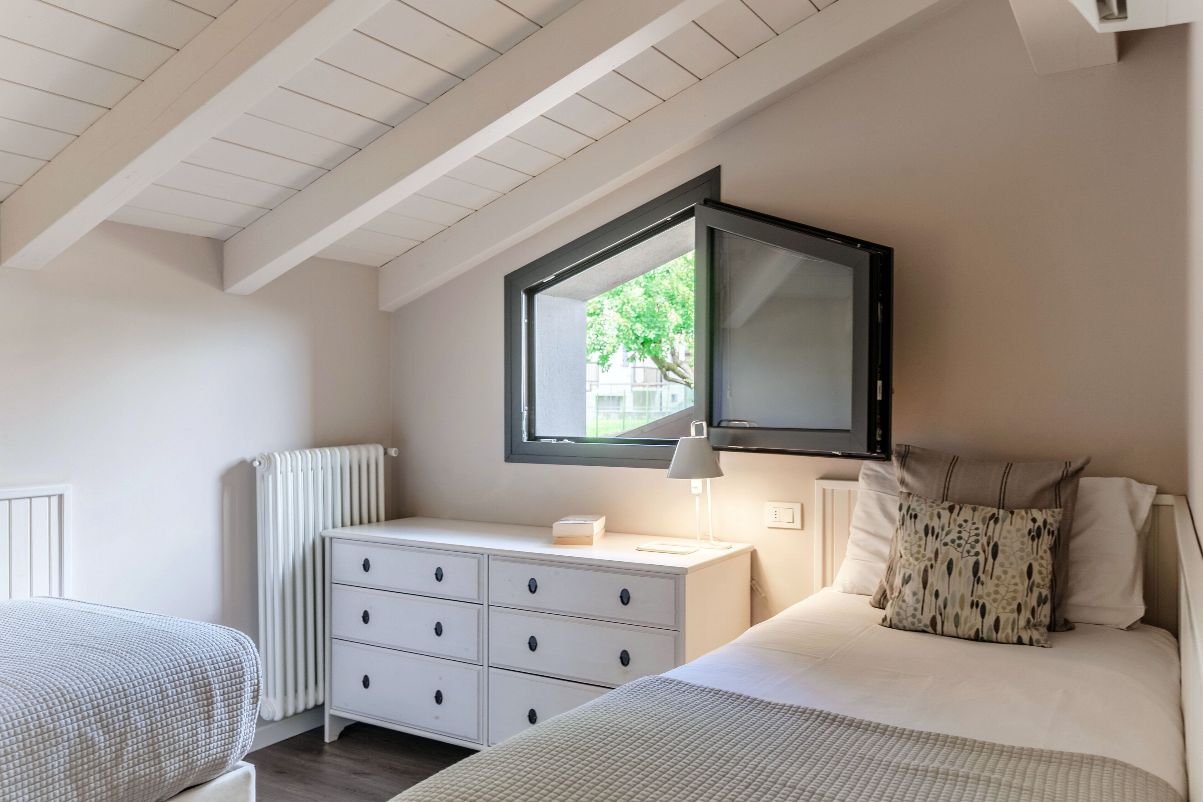 A bedroom with a bed, a nightstand, a dresser, a window, and a ceiling with wooden beams.