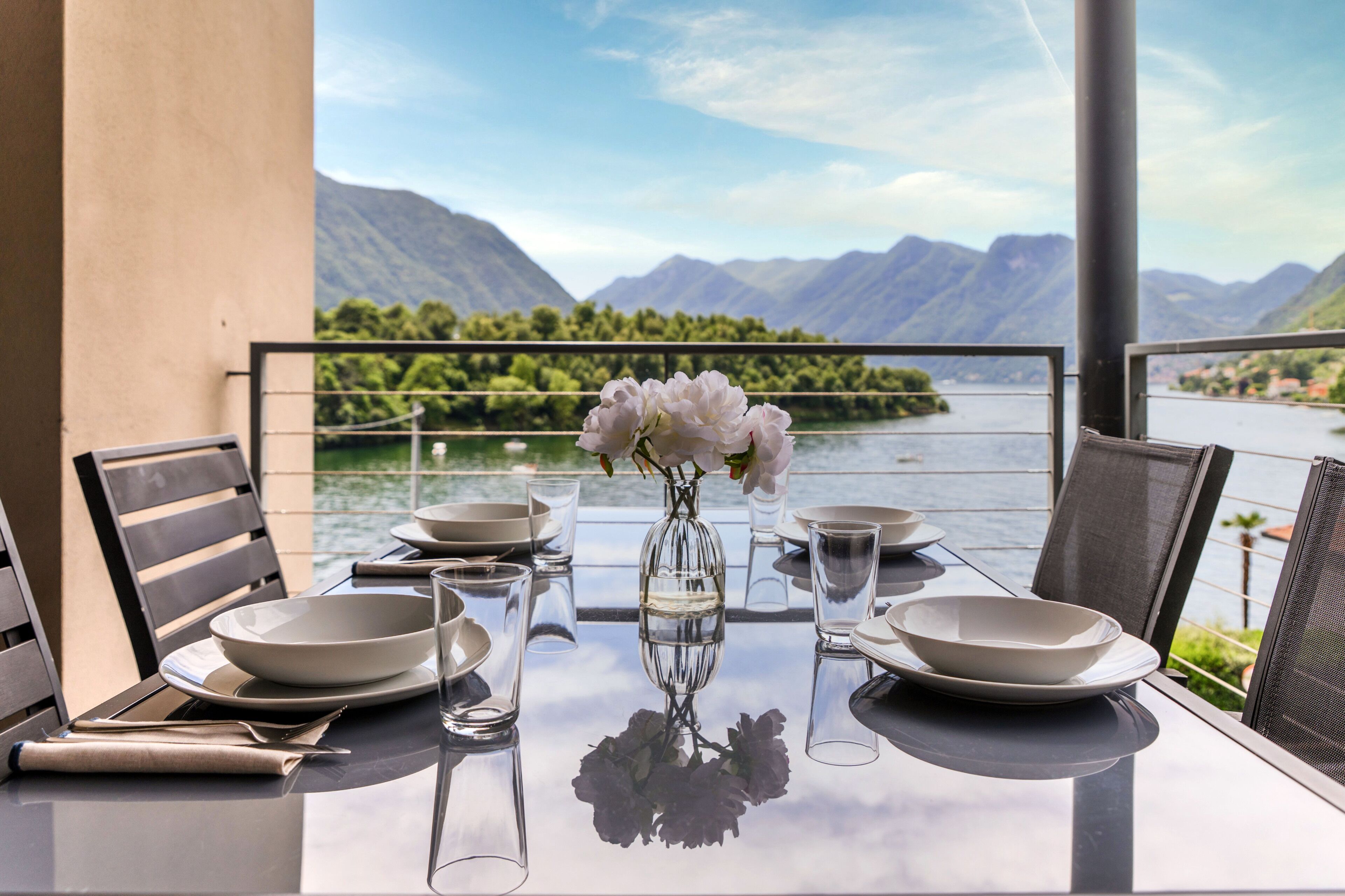 A dining table set with plates, bowls, and glasses, with a vase of flowers in the center, overlooking a lake and mountains.