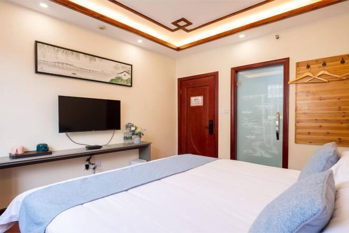 A hotel room with a bed, a flat-screen TV mounted on the wall, a wooden dresser, a desk with a computer, and a door with a wooden panel.