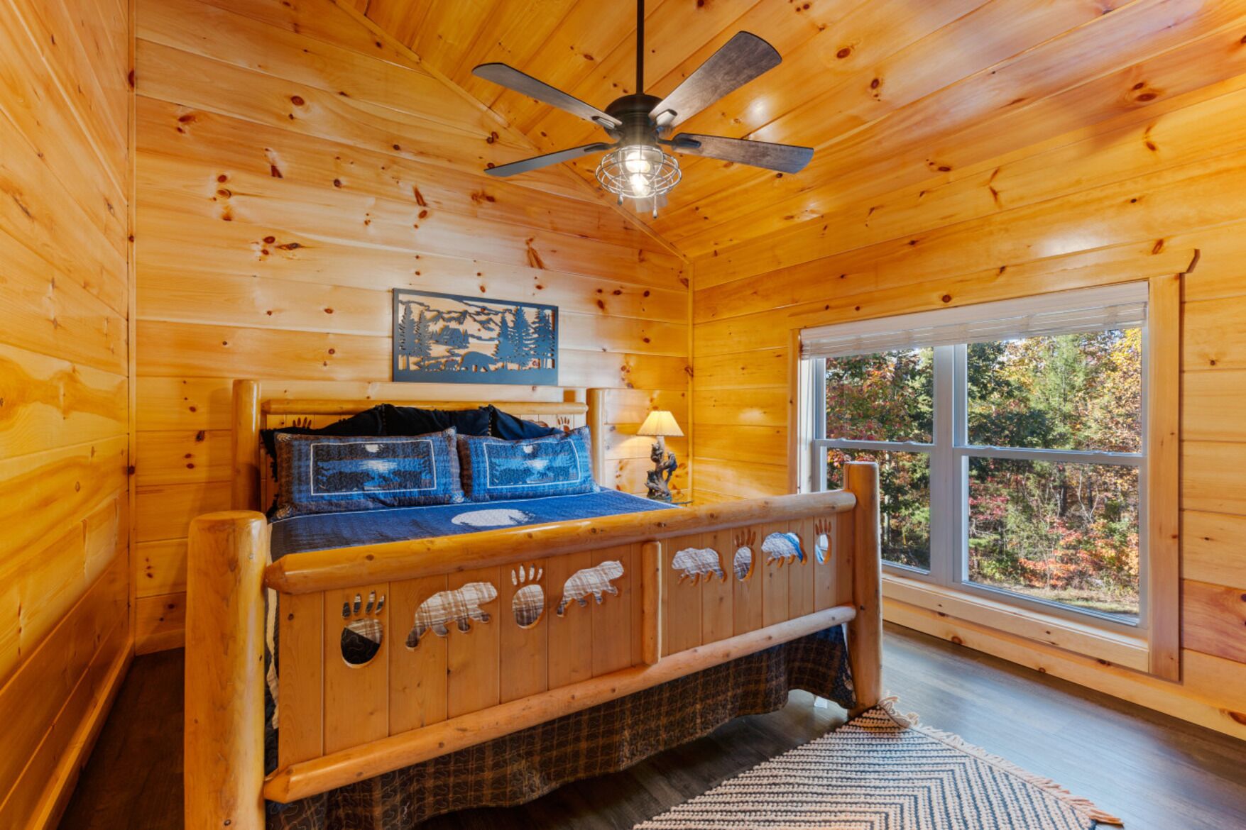 A wooden cabin bedroom with a bed, a ceiling fan, a window with a view of trees, and a wall-mounted picture.