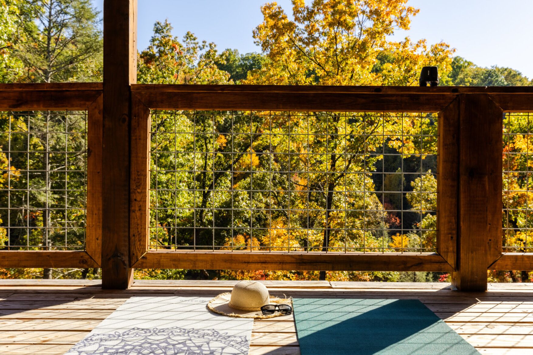 Una terraza de madera con vistas a árboles otoñales a través de una ventana con celosía metálica.