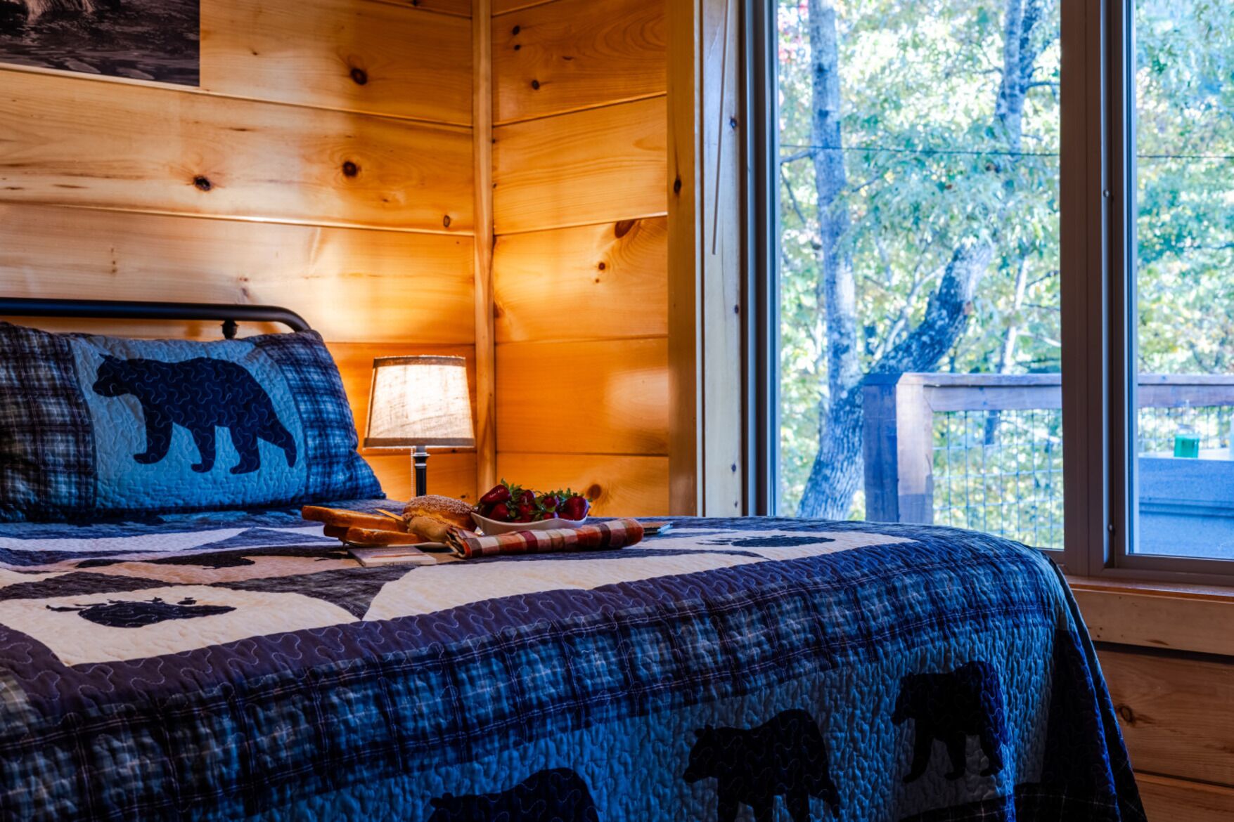 Una cama con colcha azul y negra adornada con motivos de osos, una lámpara y vista de árboles a través de una ventana.