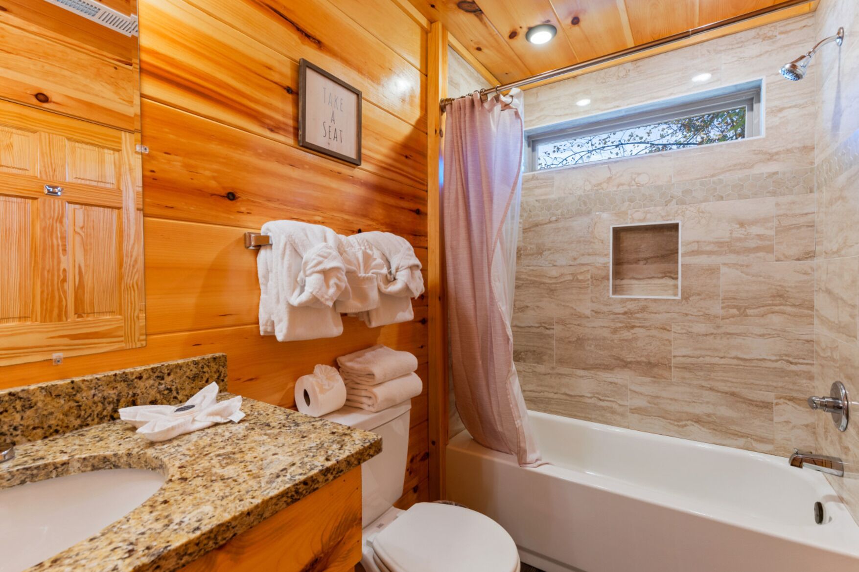 Baño de cabaña de madera con encimera de granito, inodoro blanco y ducha con pared de piedra.