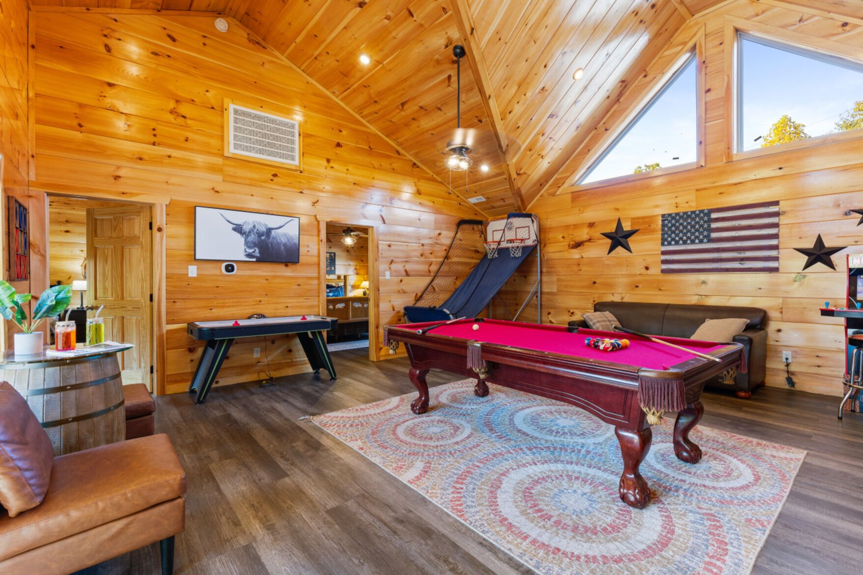 A cozy, wooden cabin interior with a pool table, a billiards table, a leather couch, and a basketball hoop.