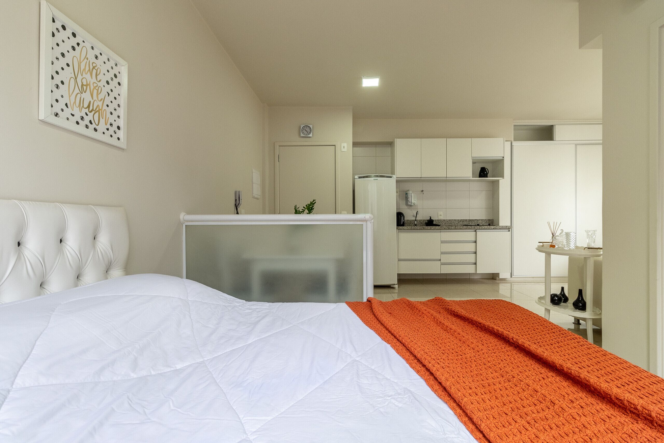 Une chambre comprenant un lit, une table de chevet, un petit coin cuisine et une salle de bain séparée par une cloison en verre.