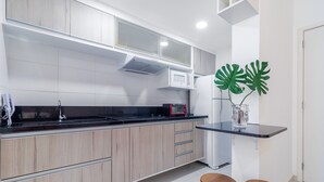 A modern kitchen with light wood cabinets, a black countertop, a microwave, and a red coffee maker. There are two wooden stools and a large green plant. - AV0905 - Studio Avanhandava (São Paulo)