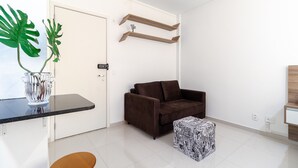 A modern living room with a brown sofa, a glass coffee table, and a patterned ottoman. There are two wooden shelves above the sofa and a door to the left. - AV0905 - Studio Avanhandava (São Paulo)