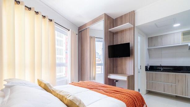 A modern hotel room with a bed, a TV, a kitchen area, and a view of the city. - AV0905 - Studio Avanhandava (São Paulo)
