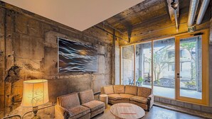 Ein geräumiger Salon mit großem Fenster, einem gemusterten Sofa, einem runden Holztisch und einer Wandlampe.