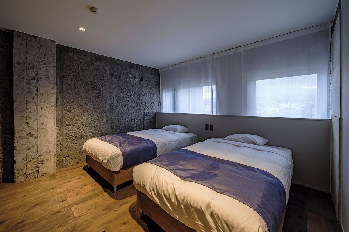 A modern bedroom with two beds, a stone wall, wooden flooring, and a large window with sheer curtains.
