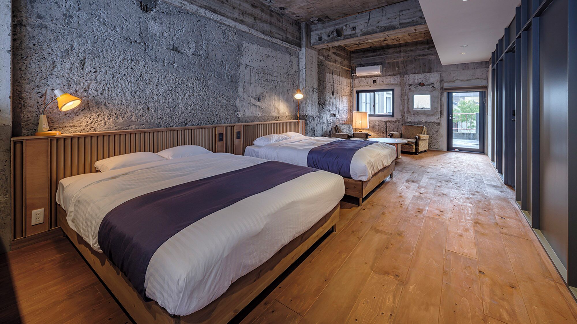 Una habitación con dos camas, suelo de madera, pared de piedra y un ventanal grande.