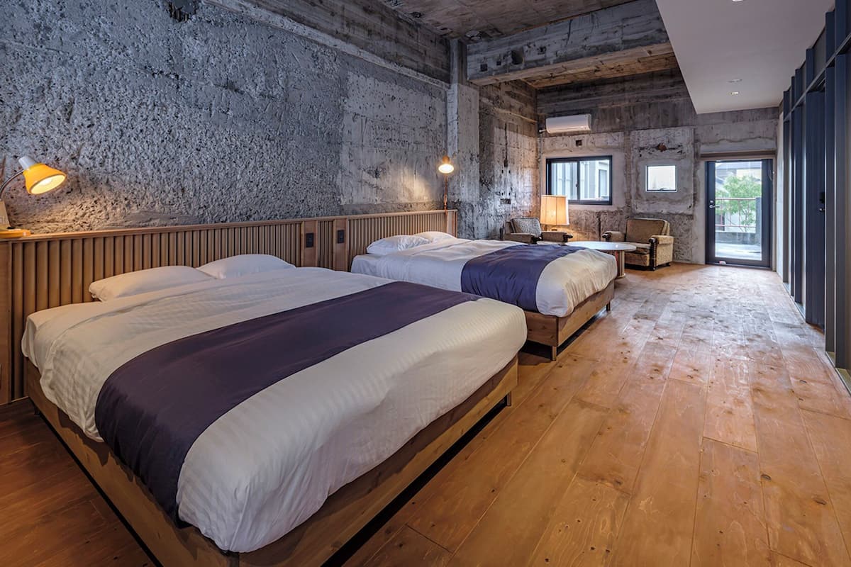 A room with two beds, wooden flooring, a stone wall, and a large window.