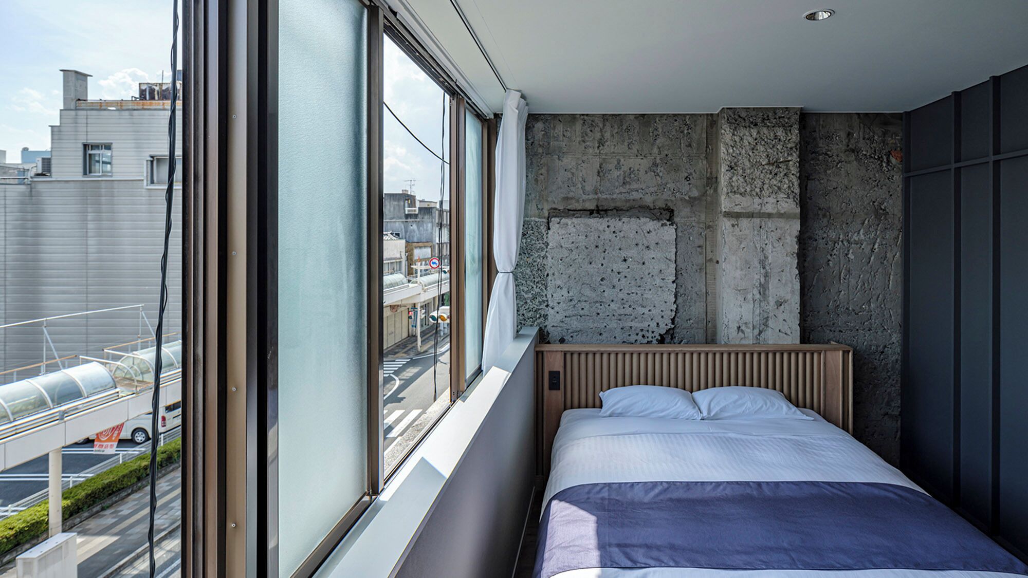 Un dormitorio con un ventanal grande, una pared de concreto y una cama.